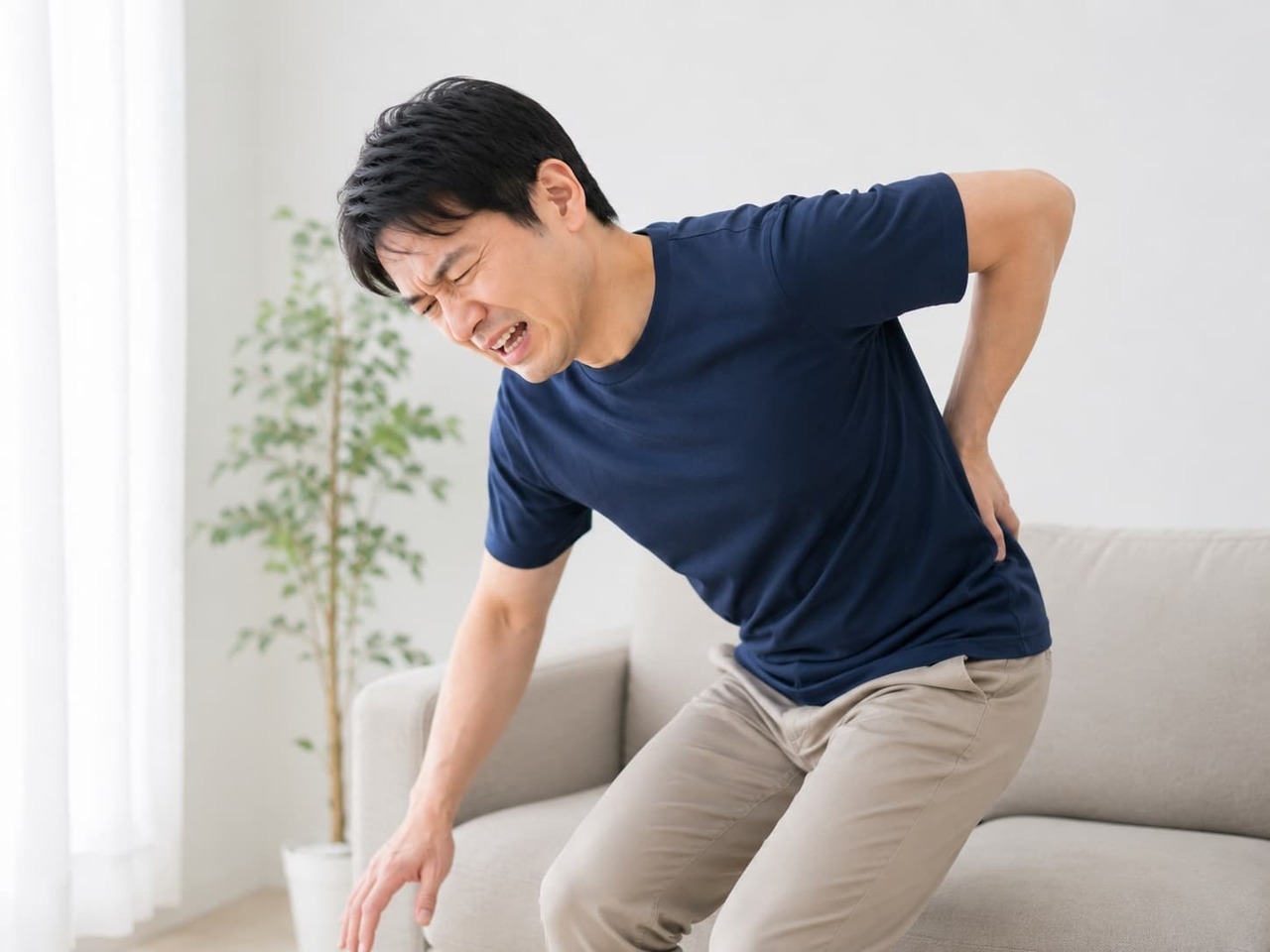 立ち上がる瞬間に腰を痛め、腰に手を当てて痛みをこらえている男性。ぎっくり腰のイメージ。